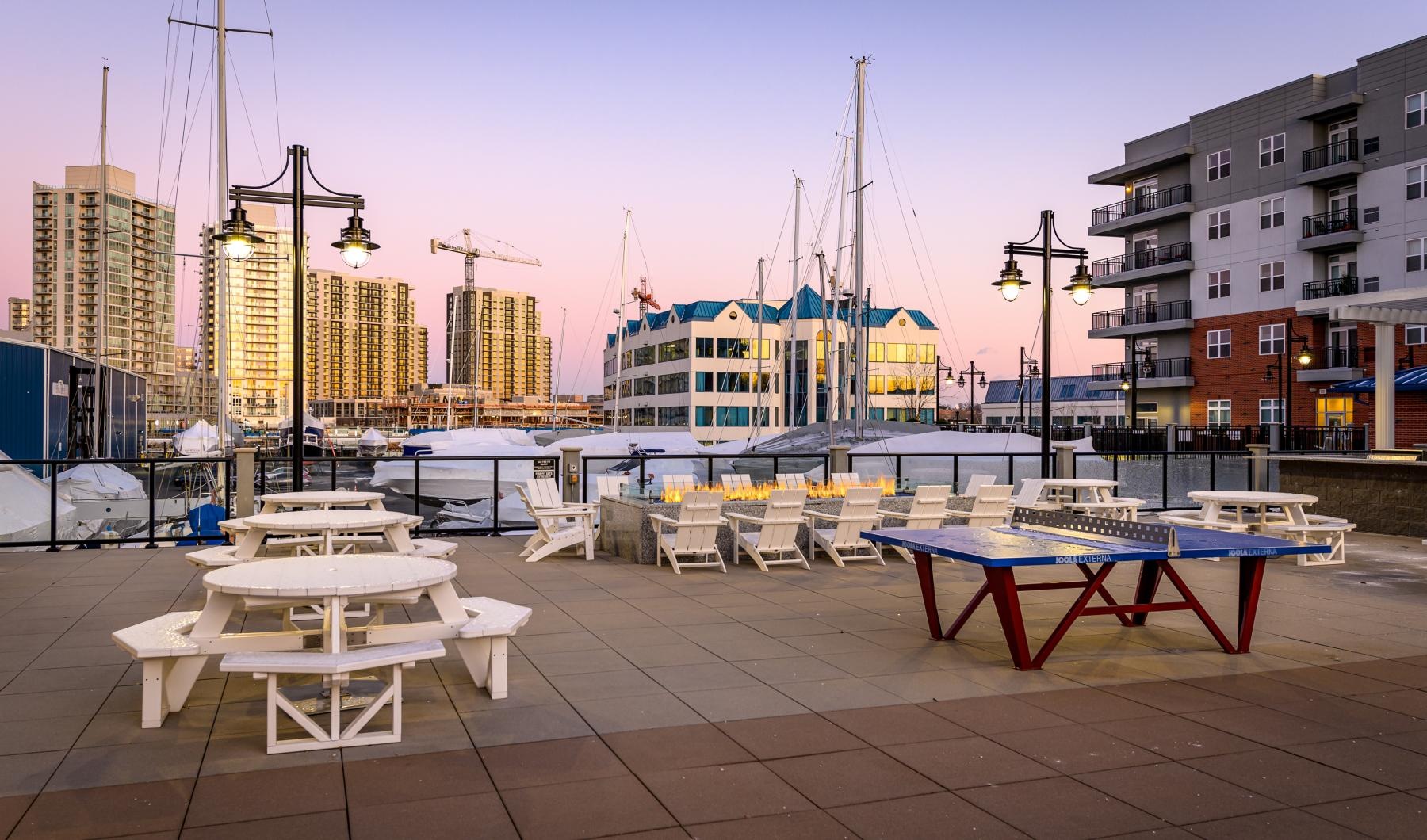 A perfect fit for the whole family Outdoor lounge at our apartments for rent in Stamford, CT, featuring adirondack chairs, a fire pit, and a view of the docks.